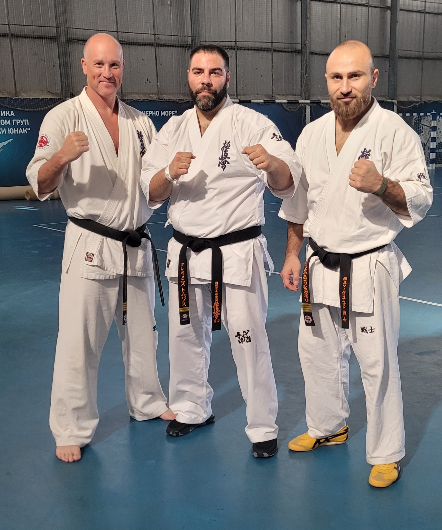 Three Kyokushin black belts standing together in a fighting stance on a blue gym floor.