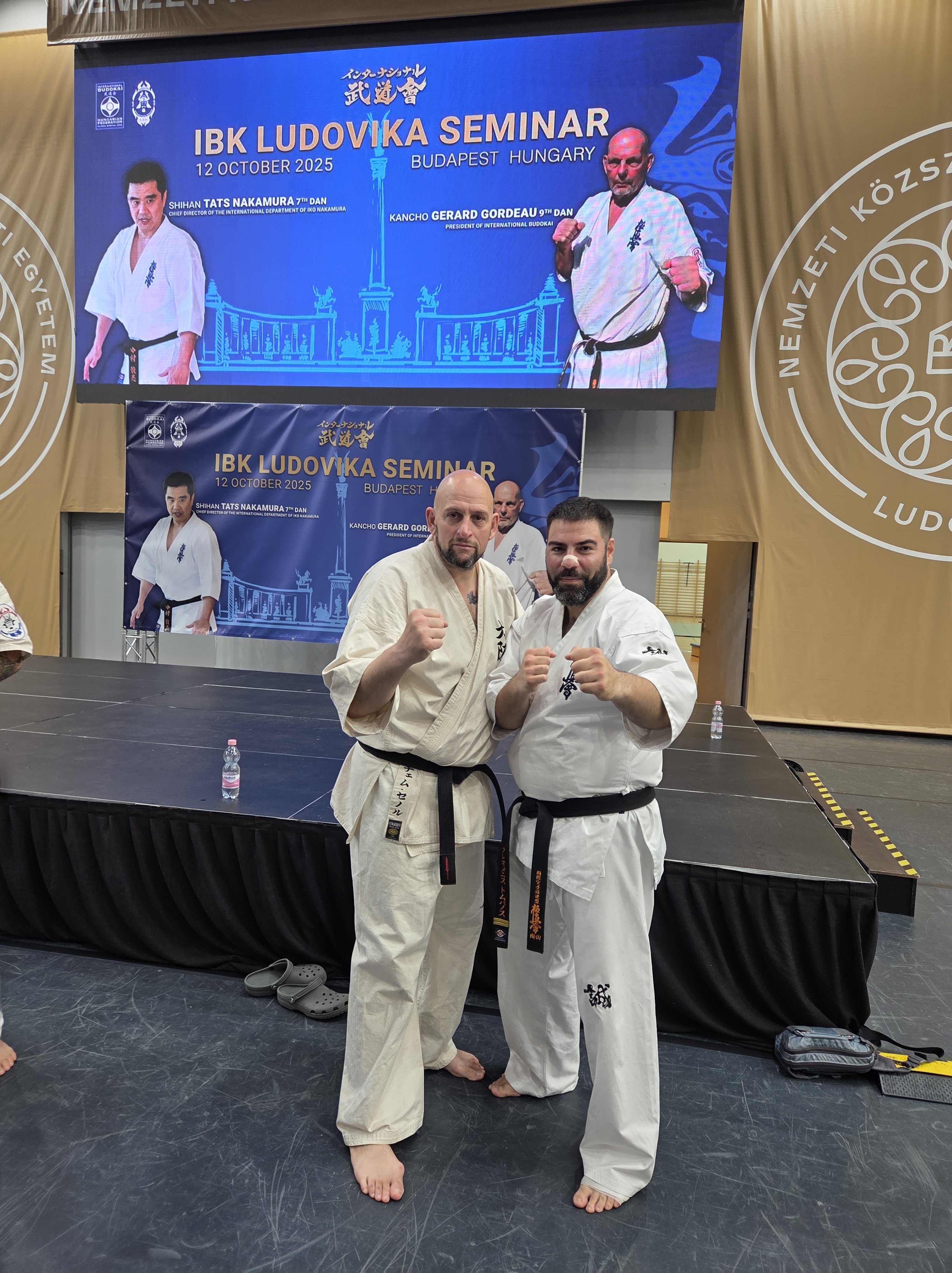 Two karate black belts posing together in fighting stances in front of a large digital banner for the IBK Ludovika Seminar 2025.