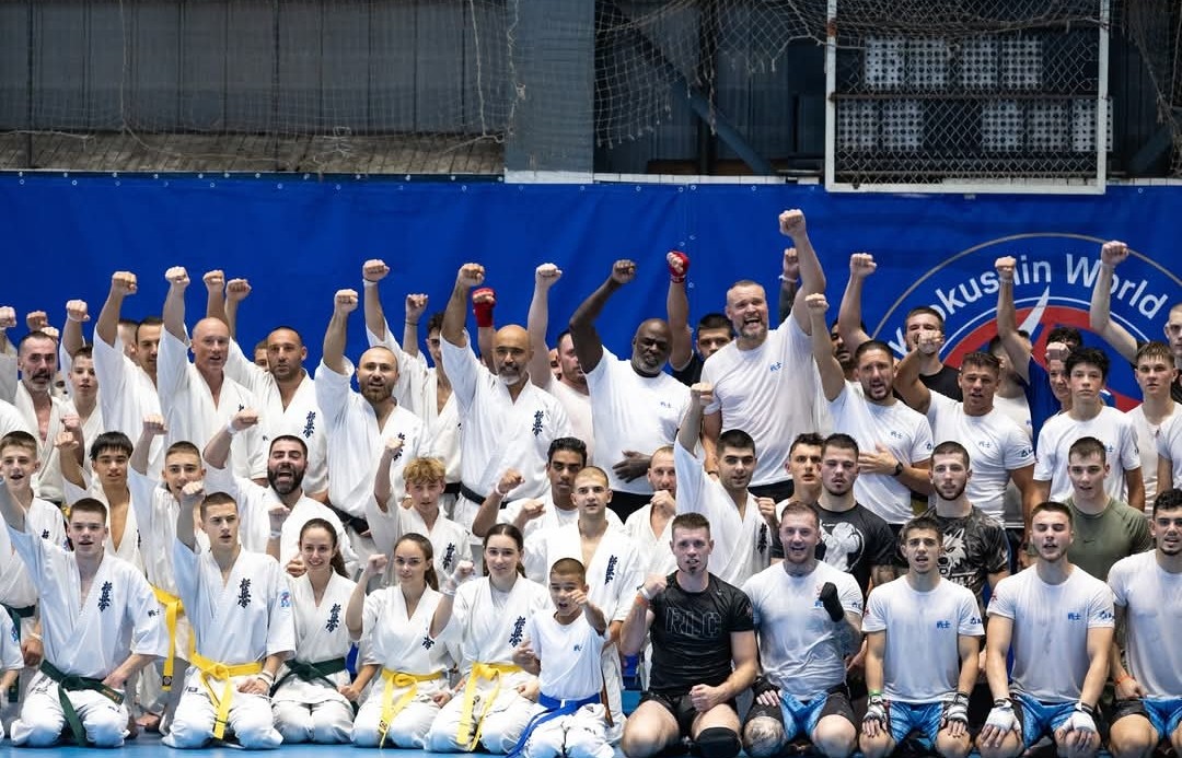 A massive group of diverse karate practitioners at Senshi Camp cheering with raised fists in front of a blue "Kyokushin World" banner.