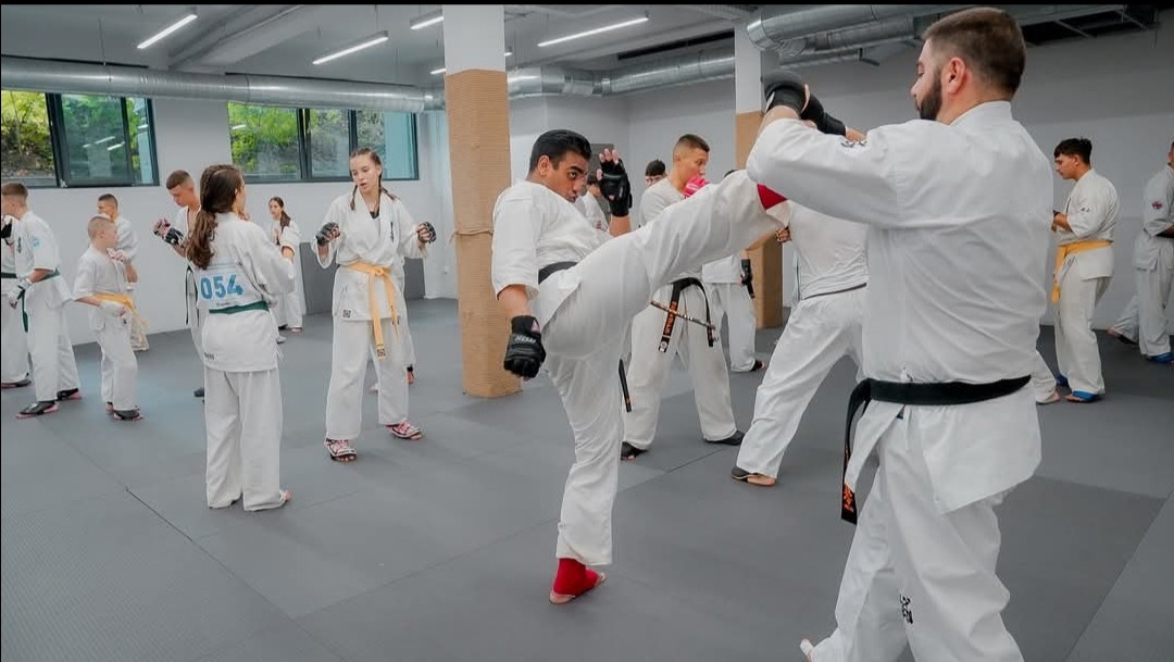 A practitioner delivering a high kick toward a black belt instructor in a modern, well-lit dojo setting.